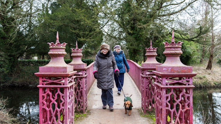 Two women walking a dog across the red bridge at Shugborough